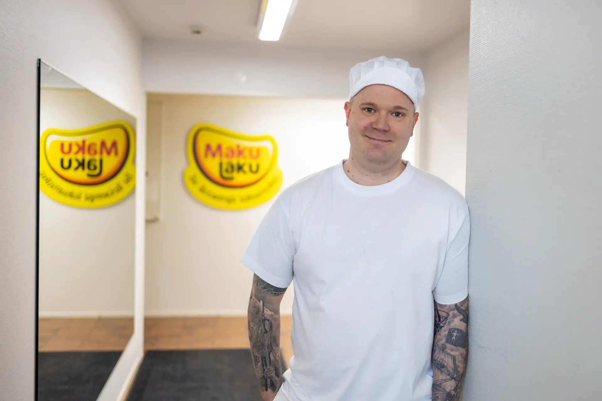 Mies valkoisessa t-paidassa ja hatussa katsoo suoraan kameraan. Taustalla seinän peittävä peili ja keltainen Maku Laku logo.