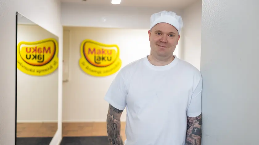 Mies valkoisessa t-paidassa ja hatussa katsoo suoraan kameraan. Taustalla seinän peittävä peili ja keltainen Maku Laku logo.