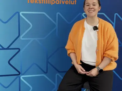 Nainen istuu jakkaralla ja katsoo suoraan eteen. Takana sininen Elis-banneri.