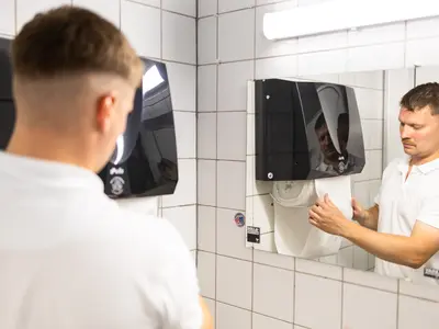 Henkilö vetää paperia seinään kiinnitetystä käsipaperiannostelijasta peilin edessä laatoitetussa pesutilassa.