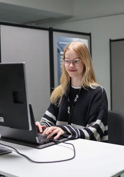 Nainen istuu työpöydän ääressä ja näpyttelee työpöydällä olevaa tietokonetta.