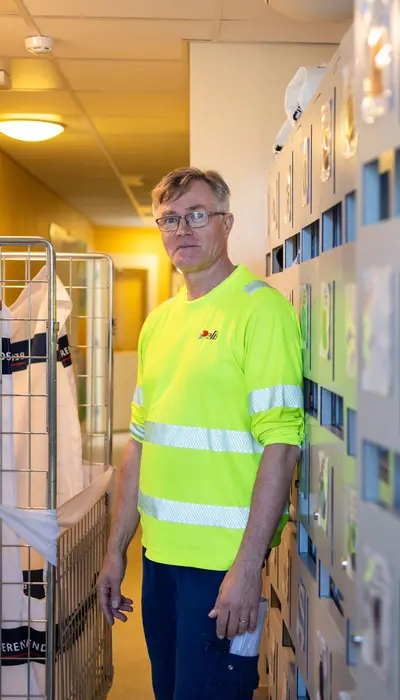 Mies nojaa hyllykköä vasten käytävällä ja katsoo kameraan.