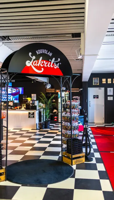 Kouvolan Lakritsin myymälän sisäänkäynti, jossa on näkyvä logo, mustavalkoinen ruutulattia, punainen matto ja lakritsituotteiden esittelytelineitä.